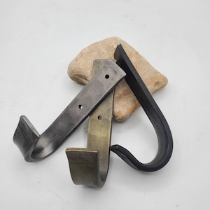 May include: Three metal hooks in varying finishes: dark gray, brass, and black. Each hook has a curved design with a rectangular base and a rounded hook. The hooks are displayed on a light-colored stone against a white background.