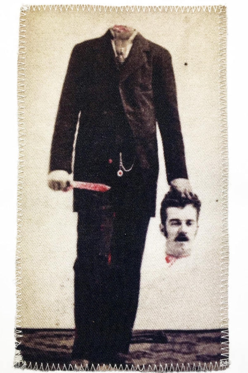 May include: A man in a suit holds a severed head in his hand. The man is holding a knife in his other hand. The background is a light beige color.