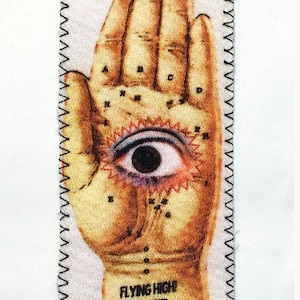 以下が含まれることがあります： 手のひらに目が描かれたヴィンテージスタイルの布製パッチ。手は文字でラベル付けされており、パッチには「Flying High」と「Lady Gonzalez」の文字が書かれています。
