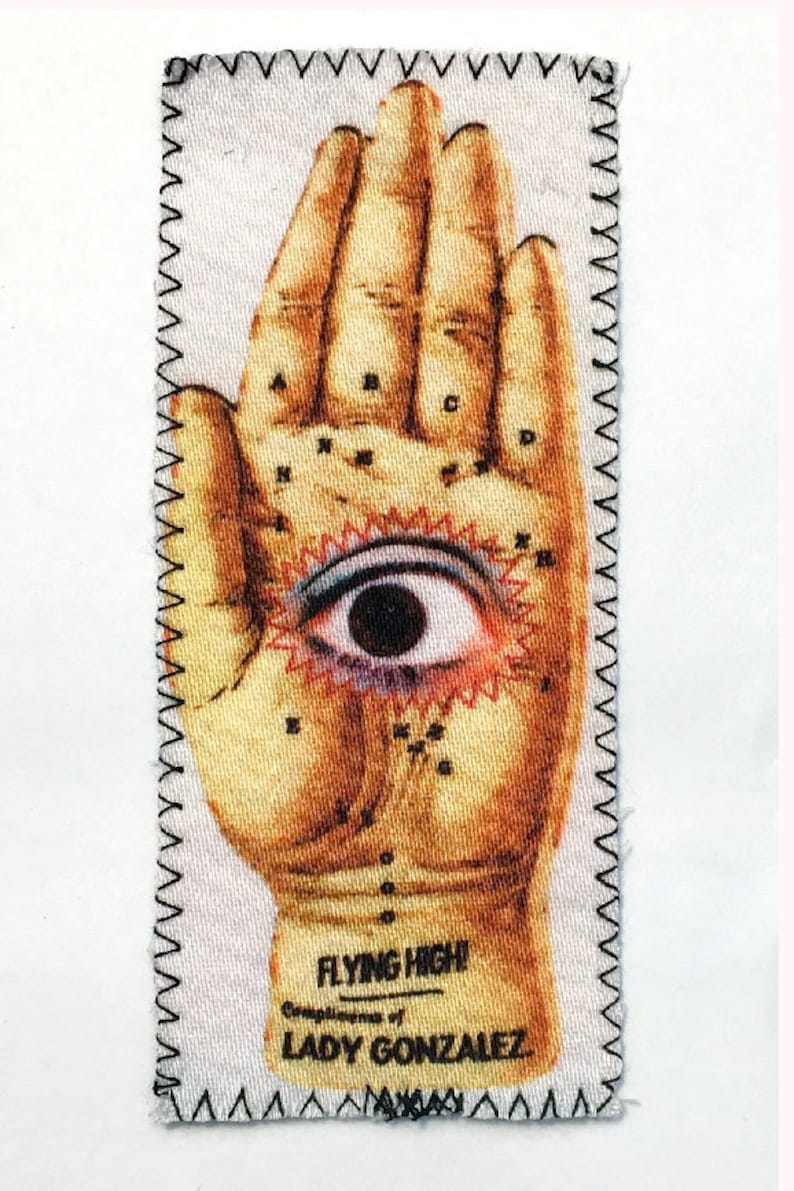May include: A vintage-style fabric patch with a hand illustration featuring an eye in the palm. The hand is labelled with letters and the patch has the text "Flying High" and "Lady Gonzalez".