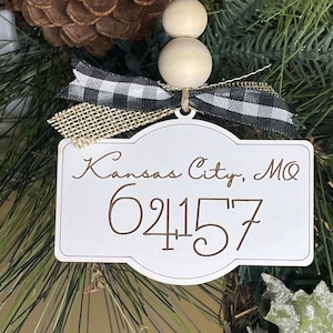 Op de afbeelding: Een witte ornament met de tekst "Kansas City, MO 64157" in bruin schrift. De ornament is versierd met een zwart-wit geruit lint, jute en houten kralen. De ornament hangt aan een groene en bruine krans.