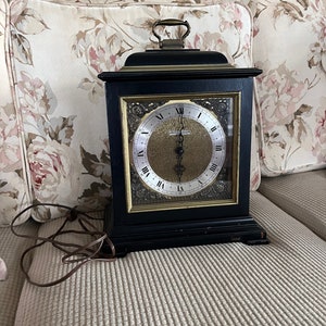 May include: A black wooden mantel clock with a gold face and Roman numeral hour markers. The clock has a brass handle on the top and is plugged in with a brown cord.