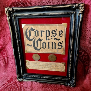 May include: A black ornate frame containing a red velvet backing with two circular metal coins. The display features a tan sign with the text Corpse Coins and a smaller sign below with text explaining the coins as payment to the Ferryman.
