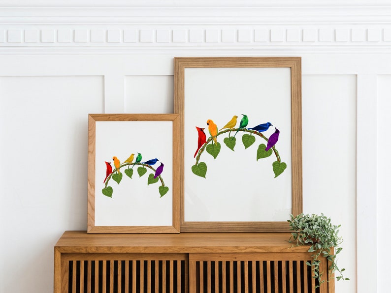 May include: Two framed art prints showing a collection of colourful birds on a leafy branch. The birds are red, orange, yellow, green, blue, and purple. The frames are light brown wood, and the artwork is displayed on a wooden cabinet.