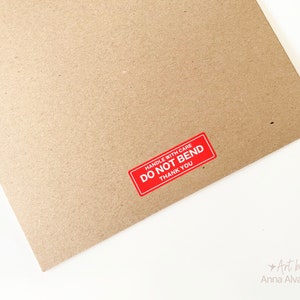 May include: A brown cardboard package with a red sticker that says "HANDLE WITH CARE DO NOT BEND THANK YOU". The package is on a white surface.