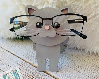 May include: Gray wooden cat figurine wearing black framed glasses. The cat is standing on a light wood surface. The cat has a pink nose and white whiskers.