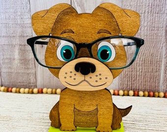 May include: A wooden dog figurine wearing glasses, with blue eyes and a brown and tan body. The dog is sitting on a green base with paw prints.