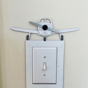 May include: A white light switch with a white toggle switch in the on position. A white airplane cutout is mounted above the switch.