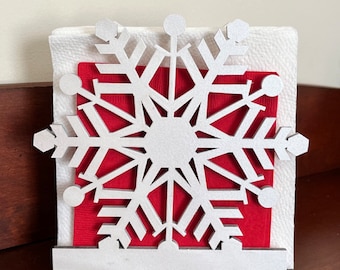 Puede incluir: Un porta servilletas de madera blanco con forma de copo de nieve con una servilleta roja en su interior. El porta servilletas tiene un acabado brillante.