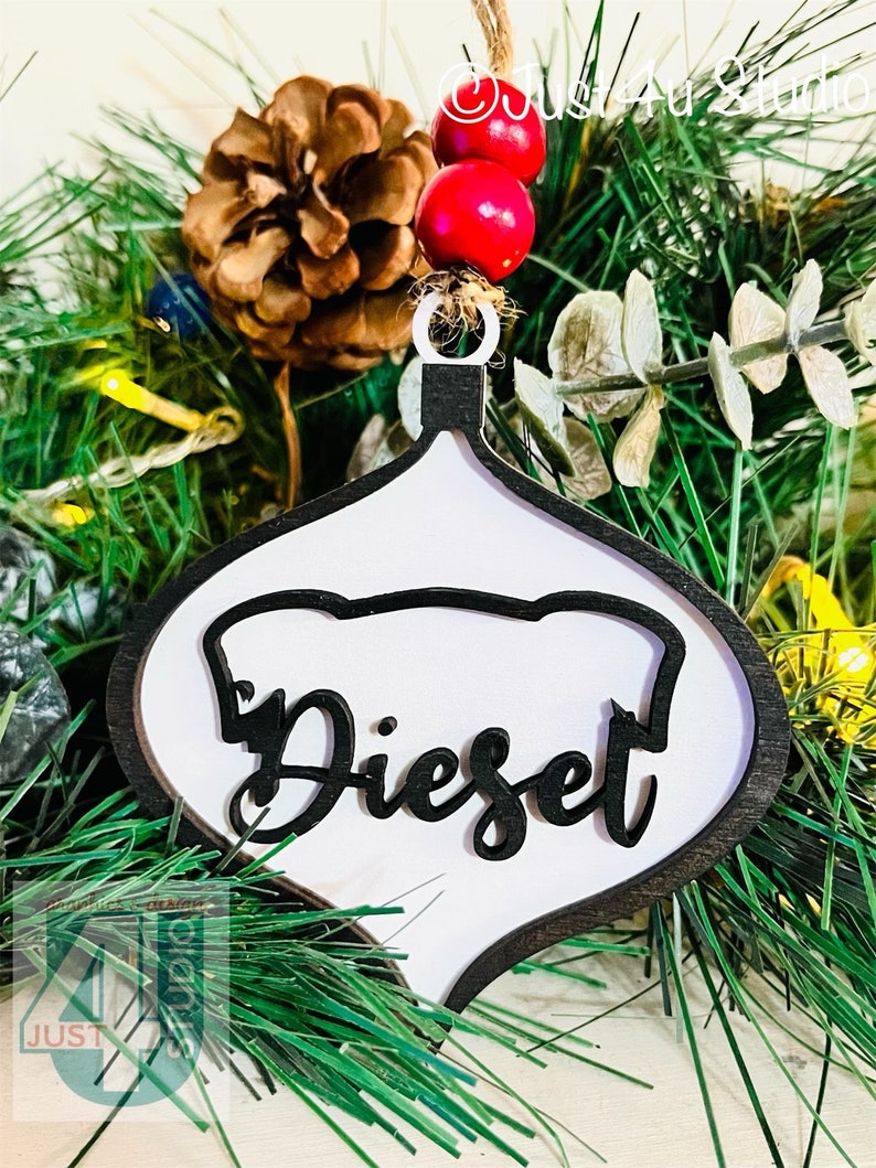 K&ouml;nnte beinhalten: Wei&szlig;er Holz-Anh&auml;nger mit schwarzem Umriss und dem Wort "Dieset" in schwarzer, kursiv geschriebener Schrift. Der Anh&auml;nger h&auml;ngt an einem gr&uuml;nen k&uuml;nstlichen Weihnachtsbaum.