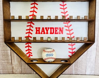 Può includere: Un ripiano in legno a forma di baseball con sfondo bianco e cuciture rosse. Il ripiano ha due livelli ed è progettato per contenere palline da baseball e altri cimeli. Il nome "HAYDEN" è stampato in lettere rosse sul ripiano. Una palla da baseball è appoggiata sul ripiano inferiore.