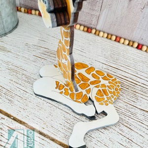 K&ouml;nnte beinhalten: Eine Holzgiraffe-Figur mit wei&szlig;em K&ouml;rper, braunen Flecken und einem langen Hals. Die Giraffe liegt, Kopf und Hals aufrecht. Die Figur hat ein detailliertes Design und ist in nat&uuml;rlichen Farben bemalt. Der Text "Just4u Studio" ist sichtbar.