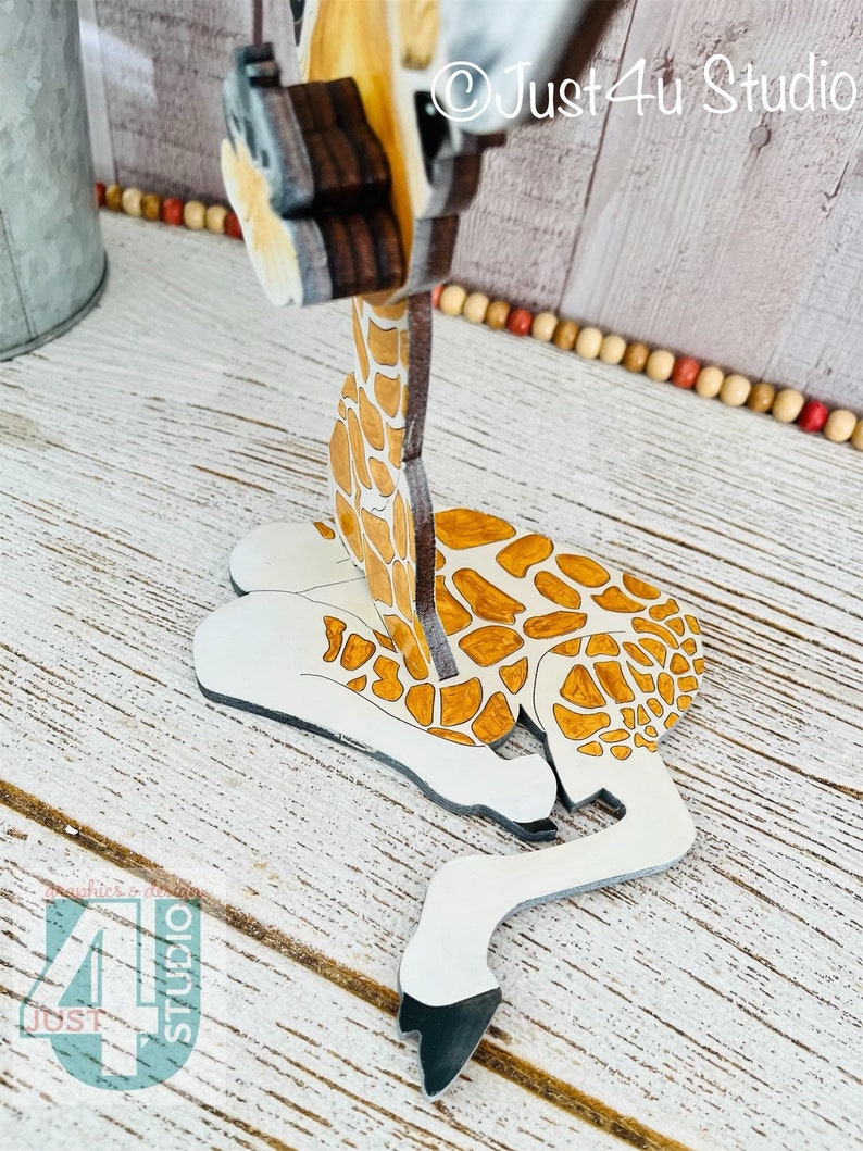K&ouml;nnte beinhalten: Eine Holzgiraffe-Figur mit wei&szlig;em K&ouml;rper, braunen Flecken und einem langen Hals. Die Giraffe liegt, Kopf und Hals aufrecht. Die Figur hat ein detailliertes Design und ist in nat&uuml;rlichen Farben bemalt. Der Text "Just4u Studio" ist sichtbar.