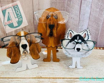 Puede incluir: Tres soportes para gafas de madera con forma de perro. El primer soporte es un basset hound marrón y blanco con gafas negras. El segundo soporte es un bloodhound marrón y blanco con gafas transparentes. El tercer soporte es un husky negro y blanco con gafas verdes. Los tres soportes están sobre una superficie de madera.