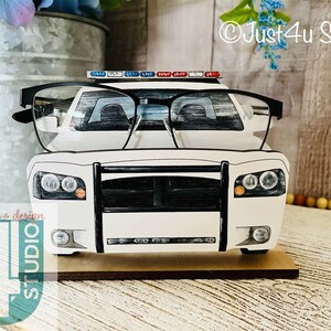 Pu&ograve; includere: Portaocchiali a forma di auto della polizia bianca con accenti neri. Il supporto &egrave; in legno e ha una piccola base. L'auto &egrave; una generica auto della polizia con una griglia e fari neri. L'auto ha una barra luminosa rossa e blu sul tetto. L'auto &egrave; appoggiata su una superficie di legno.