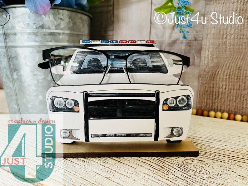 Pu&ograve; includere: Portaocchiali a forma di auto della polizia bianca con accenti neri. Il supporto &egrave; in legno e ha una piccola base. L'auto &egrave; una generica auto della polizia con una griglia e fari neri. L'auto ha una barra luminosa rossa e blu sul tetto. L'auto &egrave; appoggiata su una superficie di legno.
