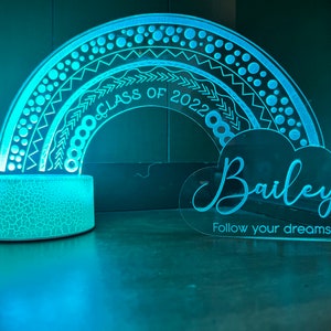 May include: A blue and white rainbow-shaped night light with a cloud-shaped plaque that reads "Bailey Follow your dreams" and "Class of 2022" on the rainbow.