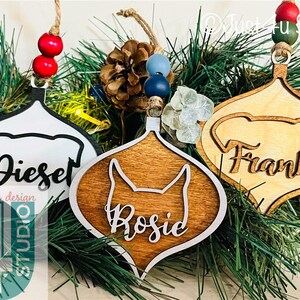 K&ouml;nnte beinhalten: Drei h&ouml;lzerne Weihnachtsbaumschmuckst&uuml;cke mit Hundesilhouetten und den Namen "Diesel", "Rosie" und "Frank" in einer kursivschriftlichen Schriftart. Die Schmuckst&uuml;cke h&auml;ngen an einem gr&uuml;nen Tannenzweig.