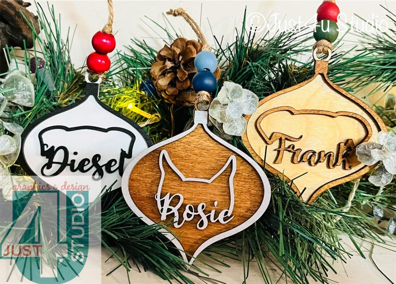 K&ouml;nnte beinhalten: Drei h&ouml;lzerne Weihnachtsbaumschmuckst&uuml;cke mit Hundesilhouetten und den Namen "Diesel", "Rosie" und "Frank" in einer kursivschriftlichen Schriftart. Die Schmuckst&uuml;cke h&auml;ngen an einem gr&uuml;nen Tannenzweig.
