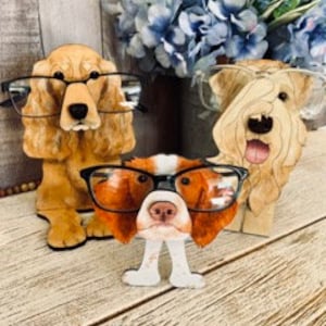 May include: Three wooden dog figurines with glasses perched on their noses. The dogs are a brown cocker spaniel, a white and brown terrier, and a brown and white spaniel. The figurines are painted in realistic detail and are a fun and unique way to display glasses.