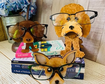 Könnte beinhalten: Drei hölzerne Brillenhalter in Form von Hunden. Die Halter sind in Brauntönen bemalt und zeigen realistische Hundegesichter mit Brillen. Die Halter werden auf einem Stapel Bücher mit dem Text "Snowed In" auf dem obersten Buch präsentiert.