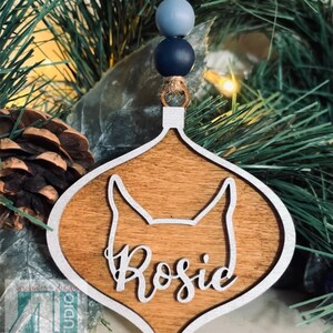 K&ouml;nnte beinhalten: Weihnachtsbaumschmuck aus Holz in Tropfenform mit silbernem Rand. Der Anh&auml;nger hat eine Hundesilhouette und den Namen "Rosie" in kursivschrift.