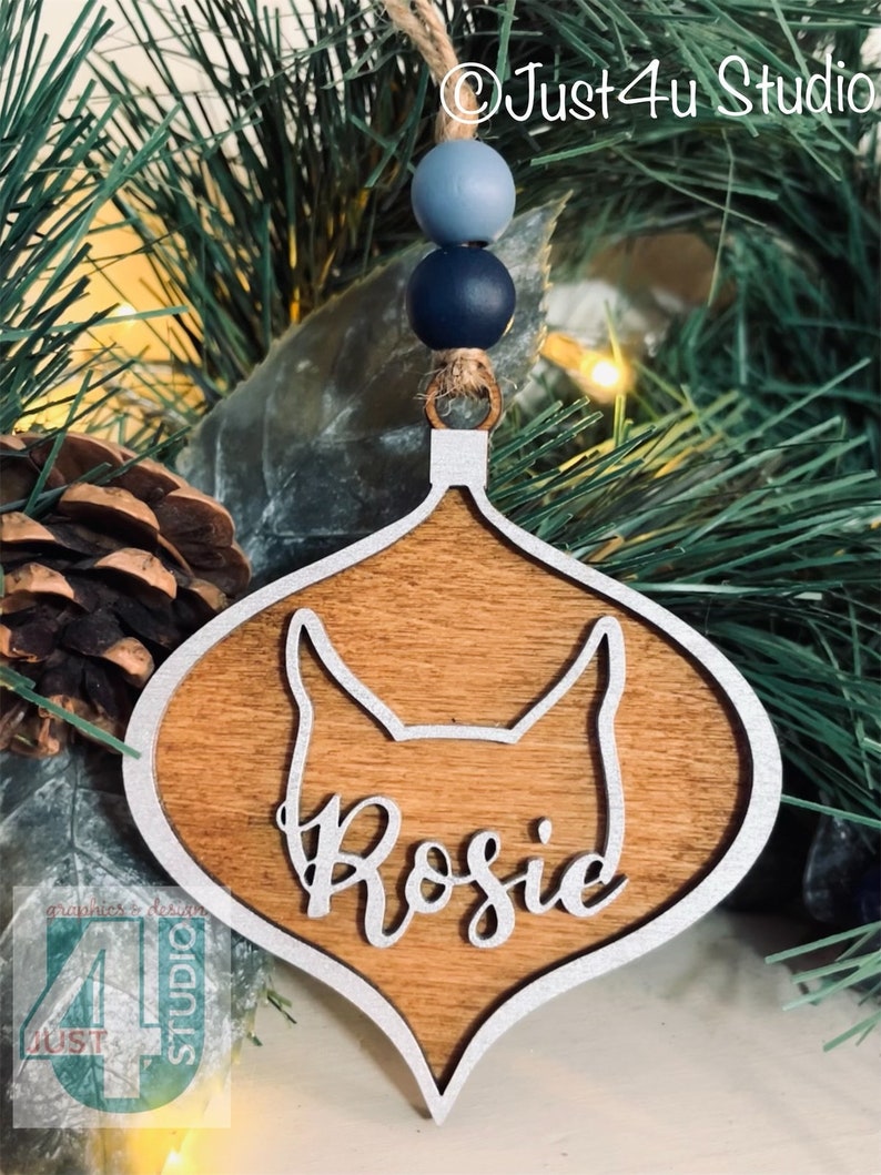 K&ouml;nnte beinhalten: Weihnachtsbaumschmuck aus Holz in Tropfenform mit silbernem Rand. Der Anh&auml;nger hat eine Hundesilhouette und den Namen "Rosie" in kursivschrift.