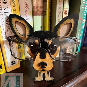 Puede incluir: Una figurita de madera de un perro con gafas de montura marrón. El perro es marrón, negro y blanco con la nariz negra y los ojos blancos. La figurita está de pie en un estante de madera frente a una estantería.