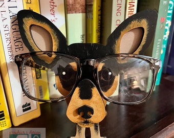 Könnte beinhalten: Eine Holzfigur eines Hundes, der eine braune Brille trägt. Der Hund ist braun, schwarz und weiß mit einer schwarzen Nase und weißen Augen. Die Figur steht auf einem Holzregal vor einem Bücherregal.