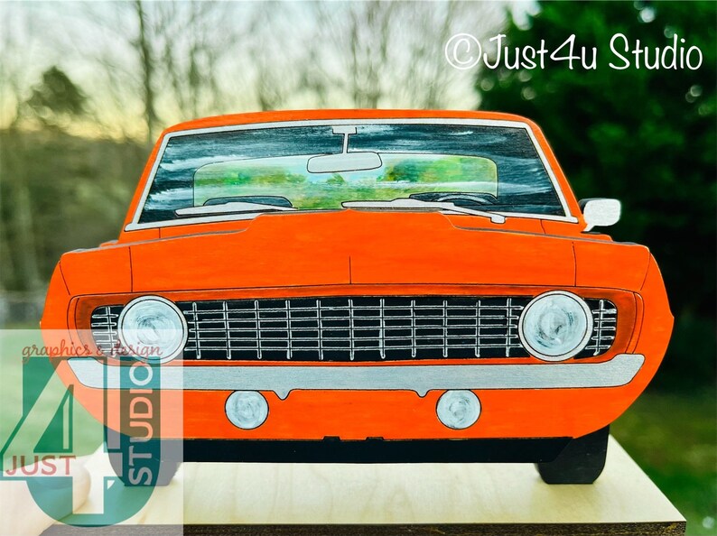 May include: A front view of a bright orange vintage car with a black grill and silver trim. The car is painted on a wooden surface.