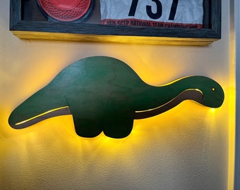 Könnte beinhalten: Eine grüne, dinosaurierförmige Nachtleuchte aus Holz, die an einer Wand montiert ist. Das Licht ist eingeschaltet und beleuchtet die Wand mit einem warmen gelben Schein.