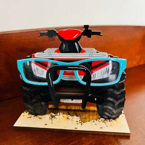 May include: A wooden toy ATV with a blue pair of glasses resting on the front. The ATV is red, black, and gray with a black metal bumper. The toy is sitting on a wooden base with a brown background.