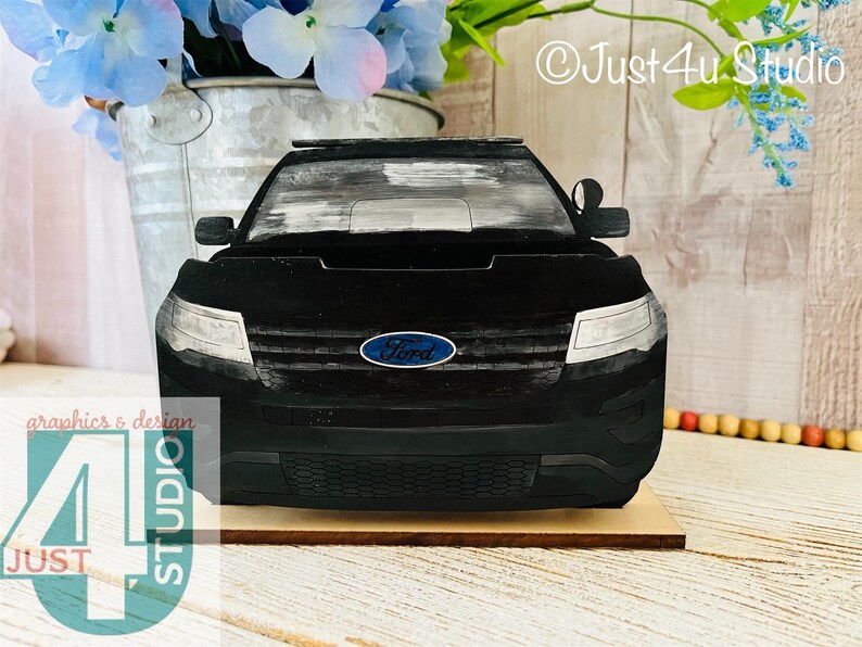 Pu&ograve; includere: Modello in legno nero di una Ford Explorer della polizia con una barra luminosa blu e bianca sul tetto. L'auto &egrave; su una base in legno. L'auto ha un logo Ford blu sul davanti.