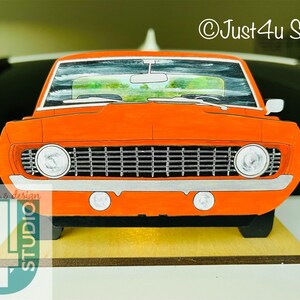 May include: Orange 1967 Chevrolet Camaro front end cutout, a wooden craft project. The car is painted orange with black accents and a white grille. The car is on a wooden base.