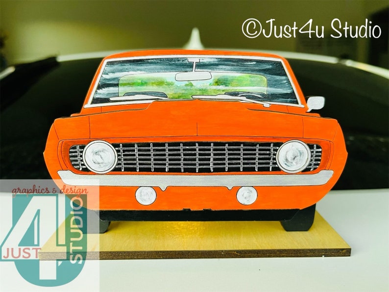 May include: Orange 1967 Chevrolet Camaro front end cutout, a wooden craft project. The car is painted orange with black accents and a white grille. The car is on a wooden base.