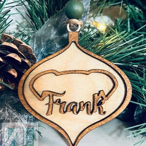 K&ouml;nnte beinhalten: Ein h&ouml;lzerner Weihnachtsbaumschmuck in Tropfenform mit einem Herz-Ausschnitt. Der Schmuck ist in einer hellen Holzfarbe bemalt und tr&auml;gt den Namen "Frank" in einer kursiv geschriebenen Schrift.