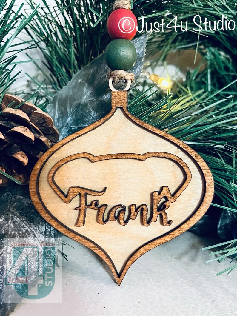 K&ouml;nnte beinhalten: Ein h&ouml;lzerner Weihnachtsbaumschmuck in Tropfenform mit einem Herz-Ausschnitt. Der Schmuck ist in einer hellen Holzfarbe bemalt und tr&auml;gt den Namen "Frank" in einer kursiv geschriebenen Schrift.