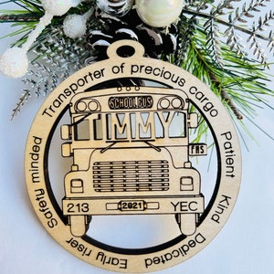 May include: A wooden ornament shaped like a school bus with the words "Transporter of precious cargo" and "Patient Kind Safety minded Dedicated Early riser" around the edge. The bus has the words "SCHOOLBUS" and "JIMMY" on the front. The bus has the number "213" and "2021" on the front.