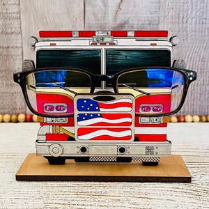 May include: A wooden fire truck-shaped glasses holder with an American flag design. The holder is painted red, white, and blue and has a black frame for the glasses.