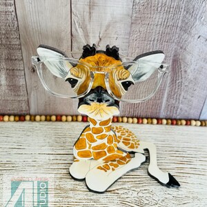 K&ouml;nnte beinhalten: Eine skurrile Giraffenfigur mit Brille mit klarem Rahmen. Die Giraffe hat ein braun-wei&szlig;es Fleckenmuster mit schwarzer Nase und Ohren. Die Figur liegt, mit dem Text "Just4u Studio" unten links.