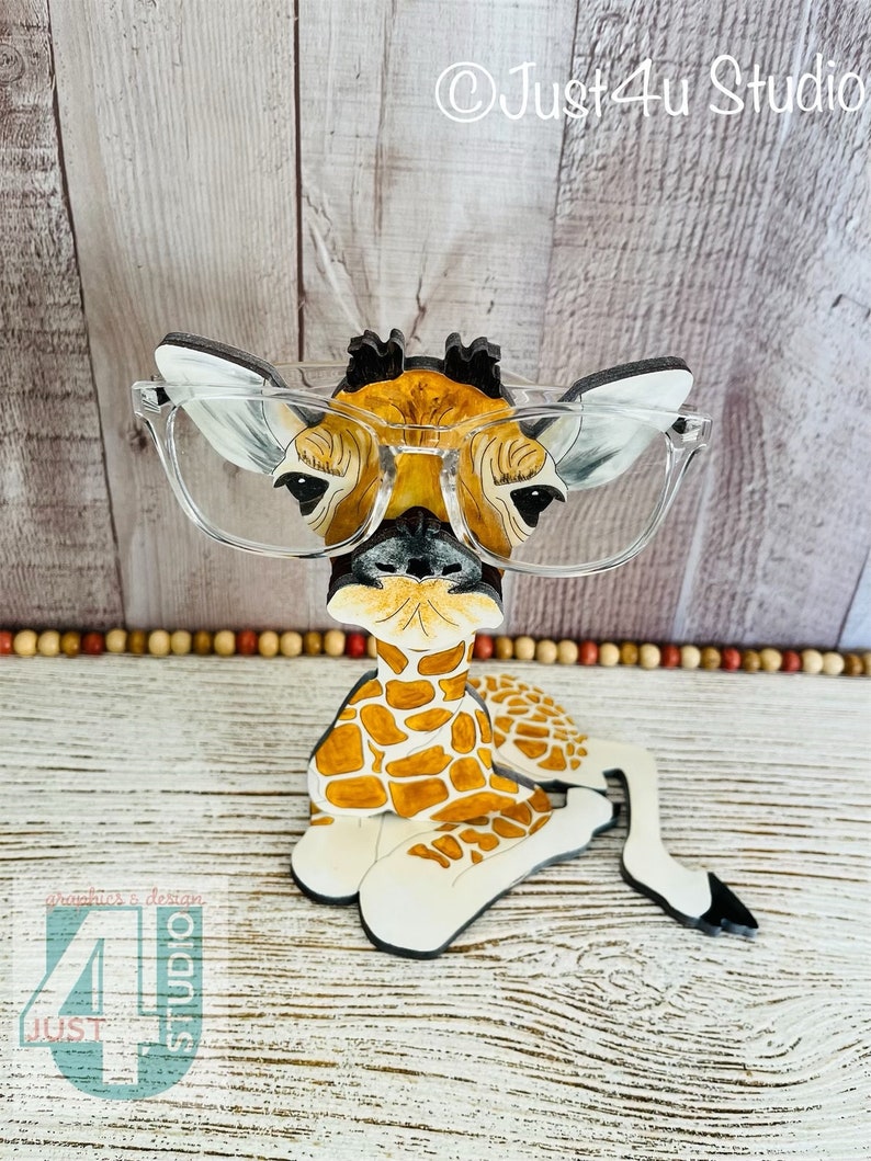 K&ouml;nnte beinhalten: Eine skurrile Giraffenfigur mit Brille mit klarem Rahmen. Die Giraffe hat ein braun-wei&szlig;es Fleckenmuster mit schwarzer Nase und Ohren. Die Figur liegt, mit dem Text "Just4u Studio" unten links.