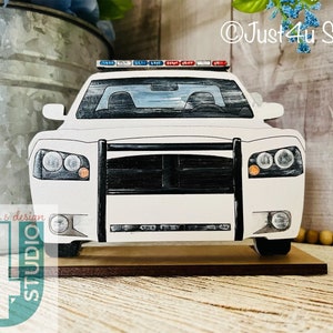Pu&ograve; includere: Taglio di auto della polizia in legno bianco con una griglia nera e luci rosse e blu in cima. L'auto &egrave; seduta su una base di legno.