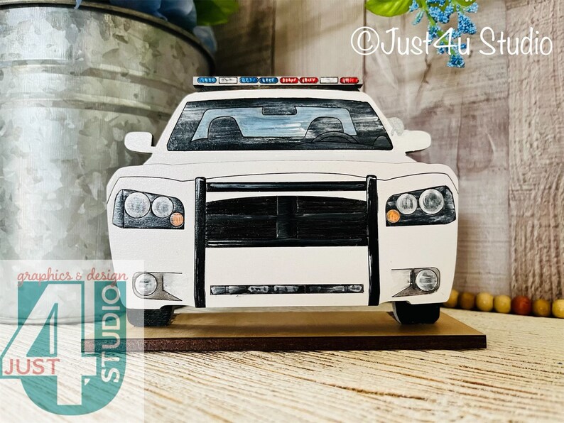 Pu&ograve; includere: Taglio di auto della polizia in legno bianco con una griglia nera e luci rosse e blu in cima. L'auto &egrave; seduta su una base di legno.