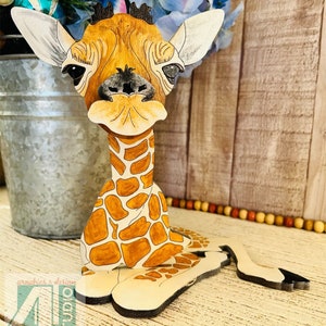 K&ouml;nnte beinhalten: Eine Holzgiraffe-Figur mit detailliertem, handbemaltem Design. Die Giraffe hat ein braunes und beigefarbenes Fleckenmuster, gro&szlig;e Ohren und einen freundlichen Ausdruck. Die Figur steht auf einer Holzoberfl&auml;che, mit dem Text "Just4u Studio".