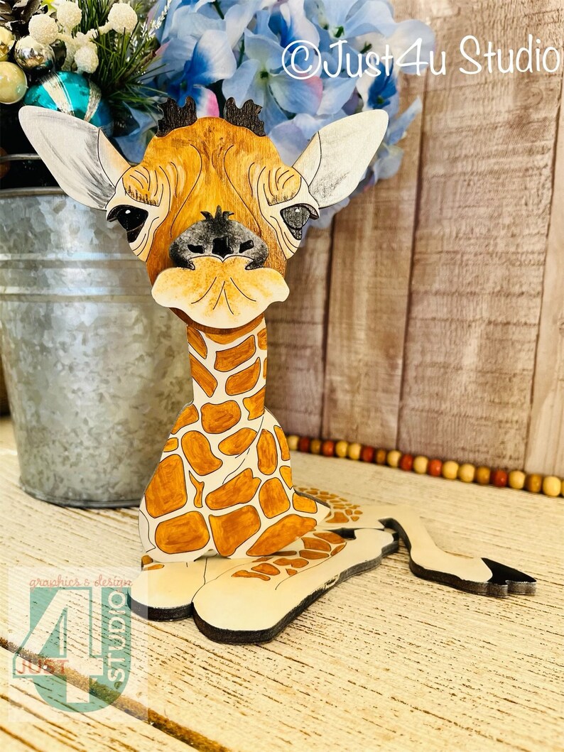 K&ouml;nnte beinhalten: Eine Holzgiraffe-Figur mit detailliertem, handbemaltem Design. Die Giraffe hat ein braunes und beigefarbenes Fleckenmuster, gro&szlig;e Ohren und einen freundlichen Ausdruck. Die Figur steht auf einer Holzoberfl&auml;che, mit dem Text "Just4u Studio".