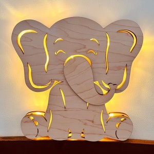 Pode incluir: Uma luz noturna de elefante de madeira com um design recortado. O elefante está sentado com as pernas cruzadas e tem um rosto sorridente. A luz está acesa e ilumina as características do elefante.