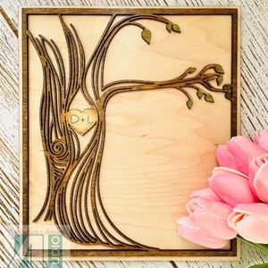 May include: A wooden cutout of a tree with a heart carved into the trunk. The heart has the initials "D+L" inside. The tree is on a light wood background with a brown frame.