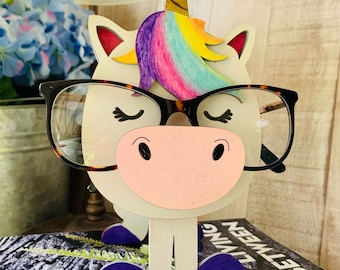 May include: A white wooden unicorn with a rainbow mane and horn, wearing glasses, and purple shoes. The unicorn is standing on a book.