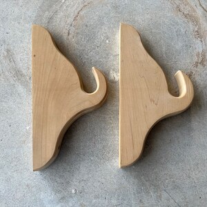 May include: Two unfinished wooden shelf brackets with a decorative curved design. The brackets have a hook on the end.