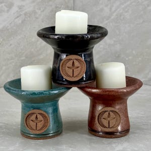 May include: Three ceramic candle holders in teal, black, and brown with a raised symbol of a flame and cross. Each holder has a white candle.